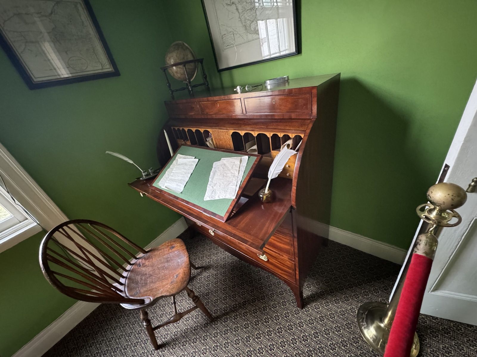 Hamilton's writing desk. Papers and a quill adorn the desk, made form oak. A single wooden chair sits empty.  A globe and artwork can also be seen in the background, against a green painted wall.
