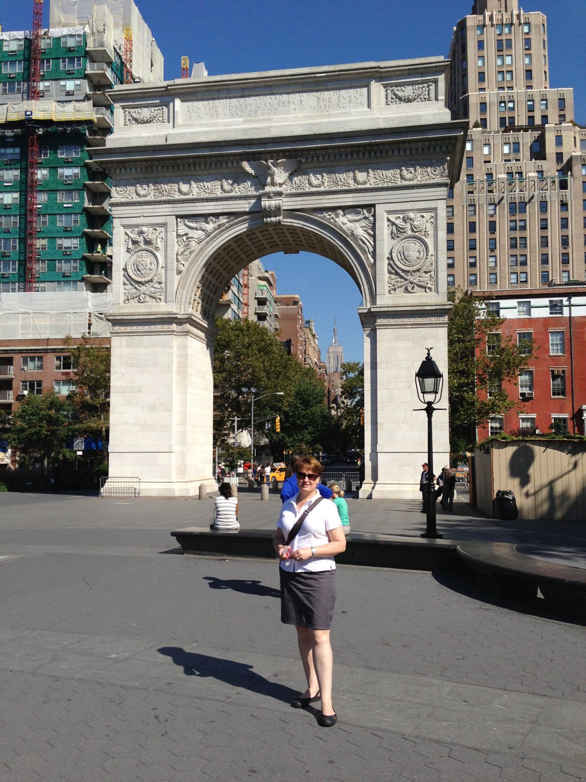 Mardi wearing a white shirt and grey skirt stands confidently in front of Washington Square Arch, showcasing her poise and the architectural design behind her. The view through the arch show 5th Avenue leading up to the Empire State Building. 