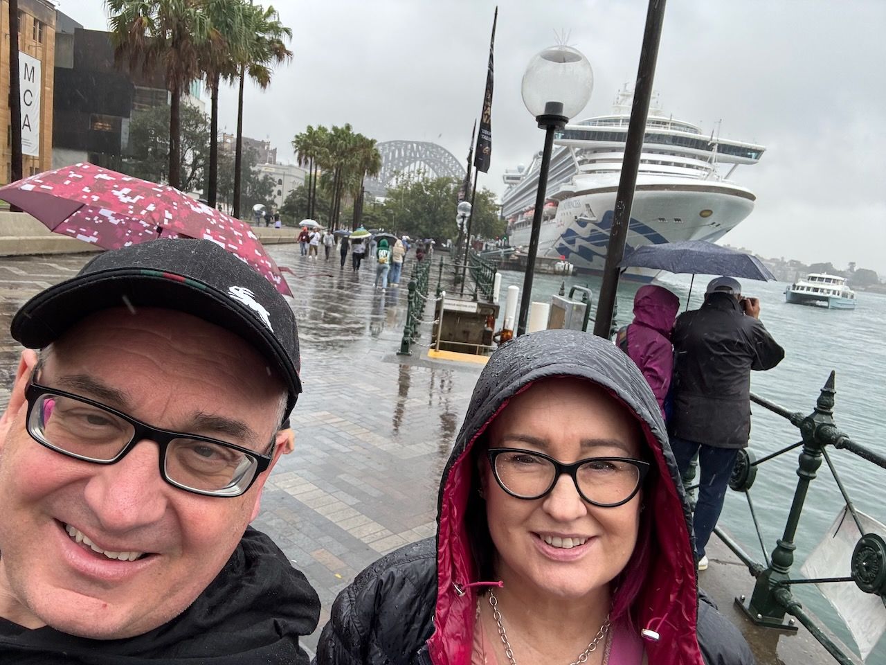 An image of michael and Marty standing at the overseas passenger terminal in Sydney it is raining very wet. The princess a large cruise ship is in the background. There are other people on the dock waiting to board as well.
